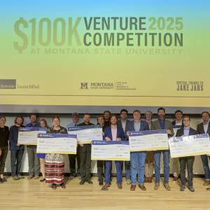 A large group stands in front of a projection screen holding large, fake checks.