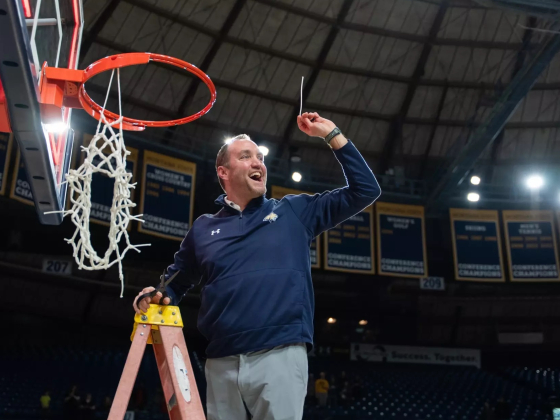 A celebratory photograph of Montana State University Athletic Director Leon Costello standing on a ladder, holding up a piece of the basketball net after a championship victory. He is wearing a navy blue MSU pullover with the Bobcat logo and has a broad s | Bobcat Athletics
