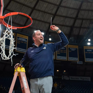 A celebratory photograph of Montana State University Athletic Director Leon Costello standing on a ladder, holding up a piece of the basketball net after a championship victory. He is wearing a navy blue MSU pullover with the Bobcat logo and has a broad s