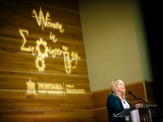 Woman speaking at podium in front of backdrop logo saying Women in Engineering.