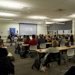 Two students stand at the front of the classroom next to a projector that says "Career Lift" with dozens of students watching them from the classroom.