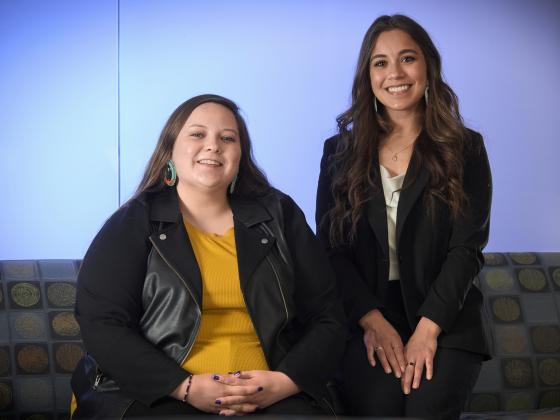 Two young women sit side by side smiling at the camera in front of a blue frosted glass background. The girl on the left is wearing hand beaded earrings and a black leather jacket over a yellow sweater. The girl on the right is wearing a black suit jacket | MSU photo by Colter Peterson