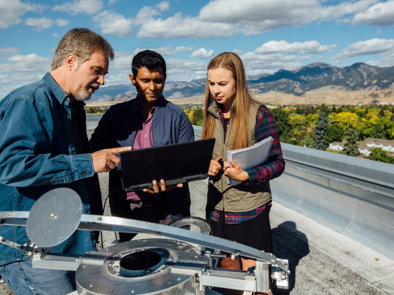 three researchers on roof looking at laptop for science with mountains in background | 