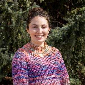 A woman with brown hair and a pink sweater smiles for a photo in front of trees