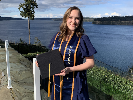 A woman stands in front of a lake holding a graduation cap with a gold tassel. She is wearing a dark blue dress and a Montana State stole. | 