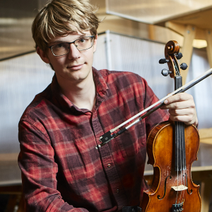 A man in a red plaid shirt holds a violin upright on his leg.