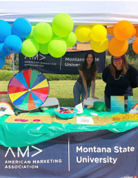 two people stand at a booth with colorful balloons, spinner, and blue banner that says American Marketing Association Montana State University