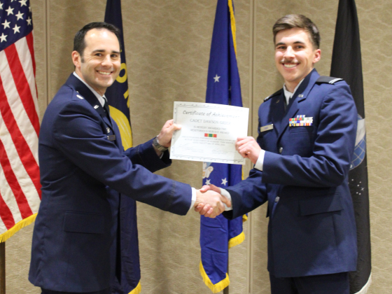 Two people wearing U.S. Air Force dress uniforms shaking hands. One is handing a certificate to another.  | 