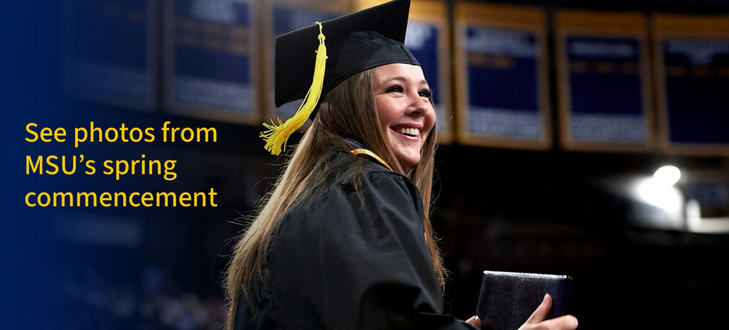 A smiling graduate in a black cap and gown with a yellow tassel holds a diploma cover while walking during Montana State University’s spring commencement ceremony. The background features championship banners hanging in an arena. Overlay text on the left  | Montana State University photo