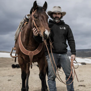 A bearded cattle rancher in a sweatshirt, black jeans and cowboy hat stands alongside a large horse in a brown leather harness.