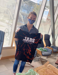 A masked person holds up a bleach dyed black t-shirt that says JABS
