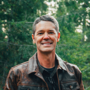 The image features a man standing outdoors in a wooded area, surrounded by lush green trees. He is wearing a brown leather jacket over a dark shirt and has a relaxed, confident expression with a slight smile. 