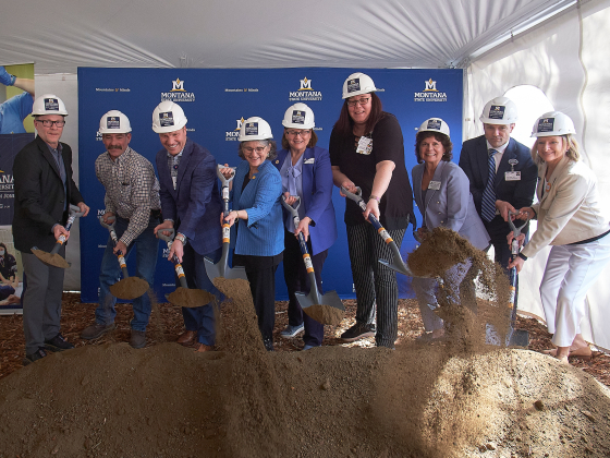 A group of people with hard hats shovel a pile of dirt during a groundbreaking ceremony. | 