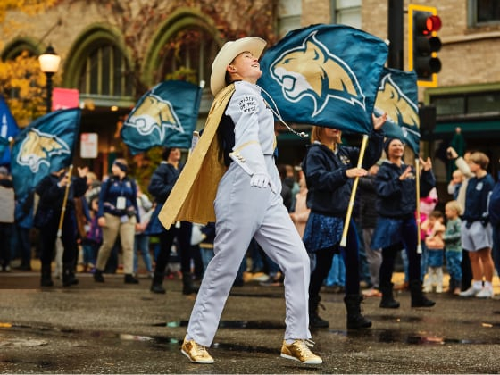A woman in a white marching band uniform strides up a street during a parade. People marching behind her carry Bobcat flags.  | MSU