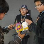 A group of three boys stand around a lego robot held by the boy of the left, appearing to discuss the design of the robot