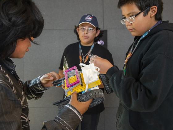A group of three boys stand around a lego robot held by the boy of the left, appearing to discuss the design of the robot | MSU photo by Colter Peterson