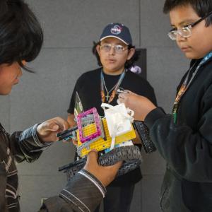 A group of three boys stand around a lego robot held by the boy of the left, appearing to discuss the design of the robot