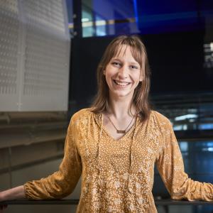 Heidi smiles while leaning against a railing wearing a yellow shirt with blue glass in the background