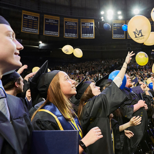 Graduates at a commencement ceremony dressed in caps and gowns watch as blue and gold balloons tumble from the rafters.