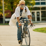 A man in a lab coat and face mask rides a bicycle on a paved path while wearing jeans.