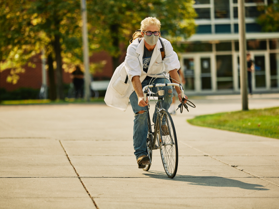 A man in a lab coat and face mask rides a bicycle on a paved path while wearing jeans. | MSU Photo by Adrian Sanchez-Gonzalez