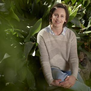 A woman sits, with her left leg crossed over the right, surrounded by green plants. She is smiling at the camera.