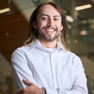 A man with long hair wearing a white collard shirt smiles at the camera in front of an out of focus staircase.