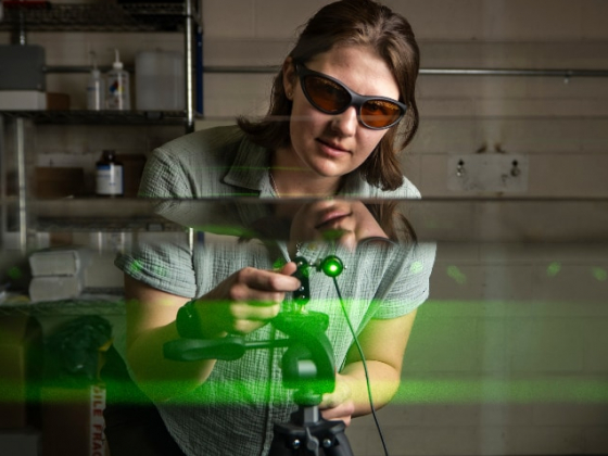 A researcher wearing protective glasses adjusts a piece of laser equipment emitting a visible green beam in a laboratory. Reflections of the beam and the researcher are seen on transparent surfaces surrounding the setup. The scene is carefully lit, highli | MSU photo by Colter Peterson