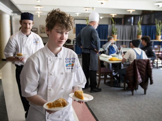 Two culinary services student swearing white chef's jackets walk single file while carrying orange desert on plates. | MSU photo by Colter Peterson