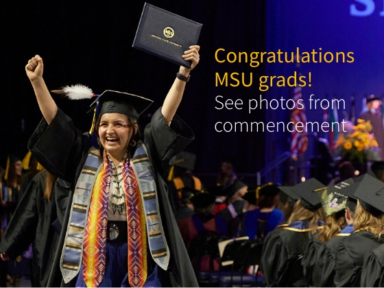A woman in graduation cap and gown holds her diploma above her head in triumph at a college commencement ceremony. | MSU