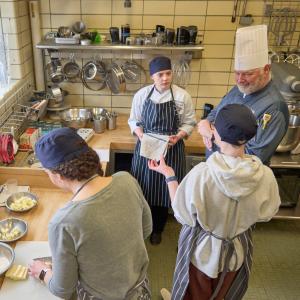 A group of people stand around a man wearing. chefs hat at a pastry station in a restaurant kitchen.