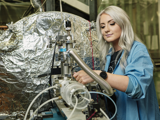 A woman in a blue lab coat makes adjustments to a large metallic scientific instrument. | MSU Photo by Adrian Sanchez-Gonzalez