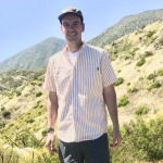 A man in a short sleeved shirt, shorts and a baseball cap stands on a sunny mountainside.