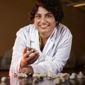 Photo of a person in a lab jacket holding volcanic rock while smiling and looking at camera.