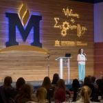 A woman stands on a stage talking to a room full of people.