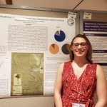 A woman in a red dress and glasses smiles in front of a research poster