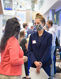 Two people in business apparel talk to each other. The student is wearing a mask and has her hair up in a bun.