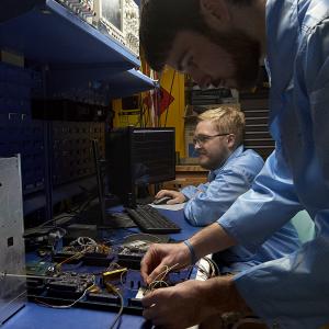 Two people wearing protective clothing work at a bench with electronics and computers