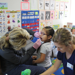 a dental hygienist examines a child's teeth in a classroom while a nursing student assists