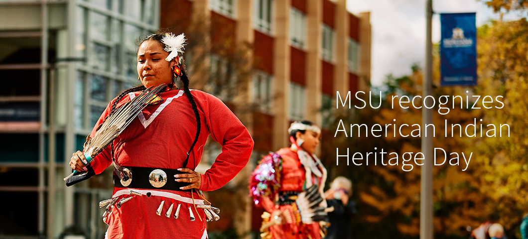 A woman in American Indian ceremonial dress in the foreground dances outdoors on a college campus in fall. | MSU