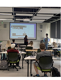 Students sitting in a classroom looking at a projector screen while a person standing at the front of the room presents.