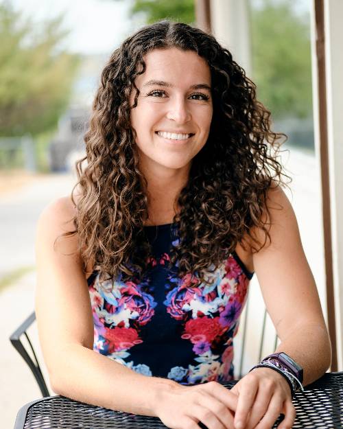 A sitting, smiling woman with long curly brown hair wearing a sleeveless floral top