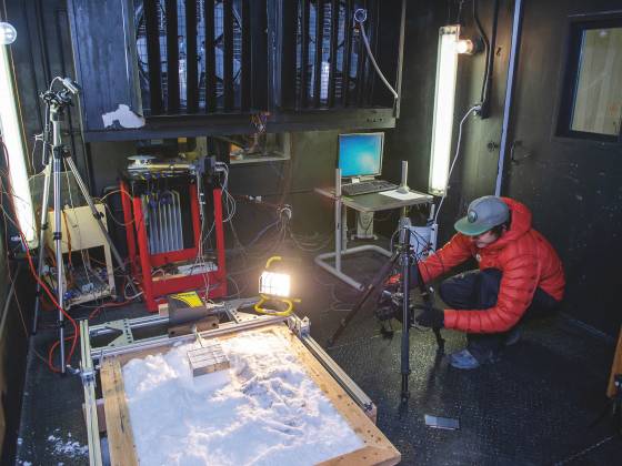 A student in red-colored down coat squats in cold room white working on device on tripod in front of pile of snow for study.