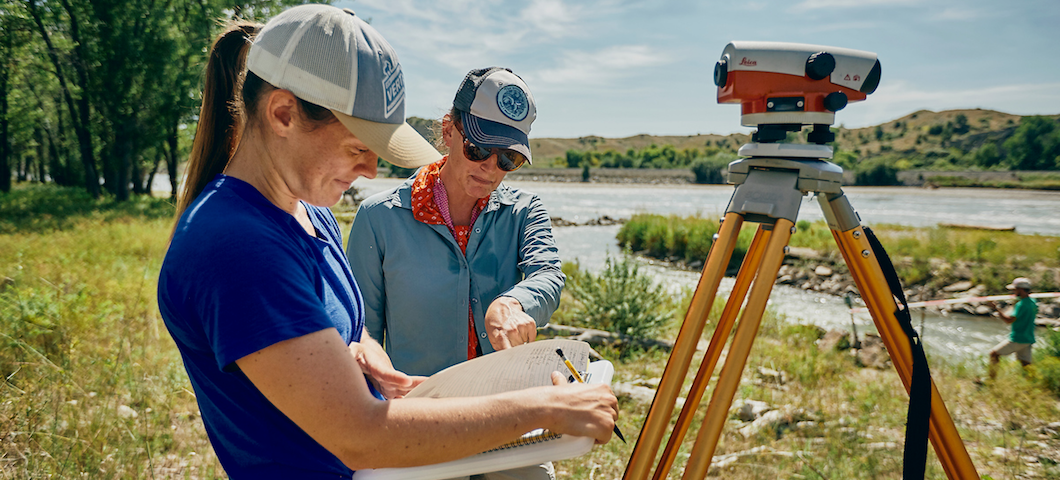 Photo of woman professor and student with surveying equipment by river and cottonwood trees in background.