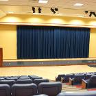 Procrastinator has tiered theater seating for 190. Lights are all the way on. Curtains are closed with a lectern. View is from the rear corner. 