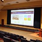 Procrastinator has tiered theater seating for 190. Overhead lights are on. Projector screen is down utilizing the ceiling mounted projector. Lectern is in the room with a spotlight. View is from side in the middle. 