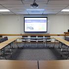 Open Square style seating. Lights are on. Projector screen is down utilizing the ceiling mounted projector. Seminar tables are the only option for tables. View is from the rear.