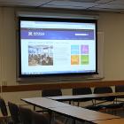 Open Square style seating. Lights are off. Projector screen is down utilizing the ceiling mounted projector. Seminar tables are the only option for tables. View is from the entry side of the room.