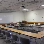 Open Square style seating. Lights are on. White board in the front with a projector screen that can be pulled down. Ceiling mounted projector. Seminar tables are the only option for tables. View is from the rear.