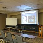 Room 233 is set with classroom style seating and a panel table. Lights are on. Projector screen is down utilizing the ceiling mounted projector. White board and lectern are up front. Air wall is up. View is from the entry door side.